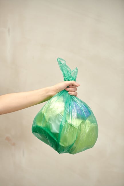 A person's arm extends into the frame from the left, holding a green plastic trash bag with a knot at the top, filled with waste. The bag appears semi-transparent, revealing various items inside, and 