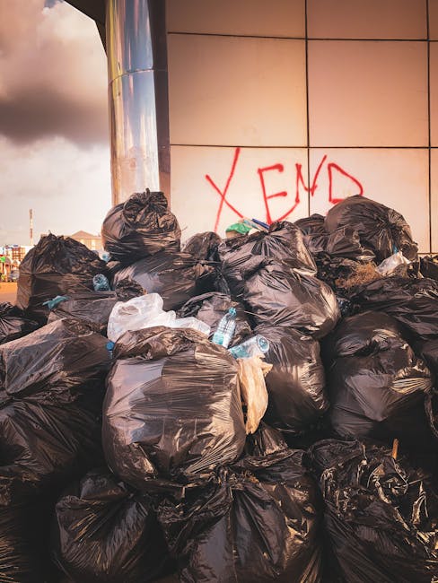 A person's arm extends into the frame from the left, holding a green plastic trash bag with a knot at the top, filled with waste. The bag appears semi-transparent, revealing various items inside, and 